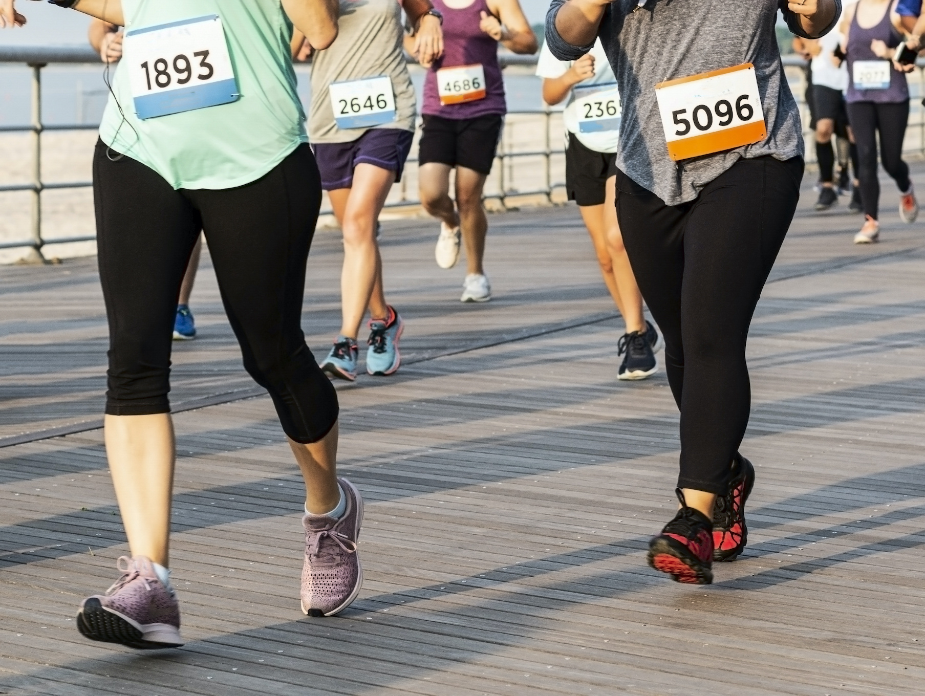 Many runners are running a race on a boardwalk by the beach shot from the waist down.
